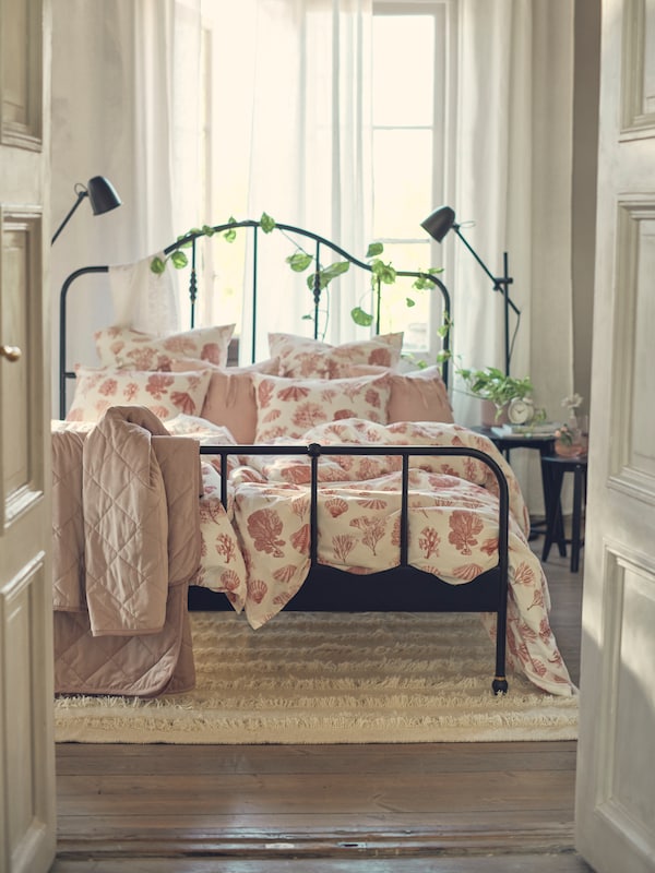 A black SAGSTUA bed with VITPYROLA bed linen stands on a PEDERSBORG rug next to a window in a bedroom.