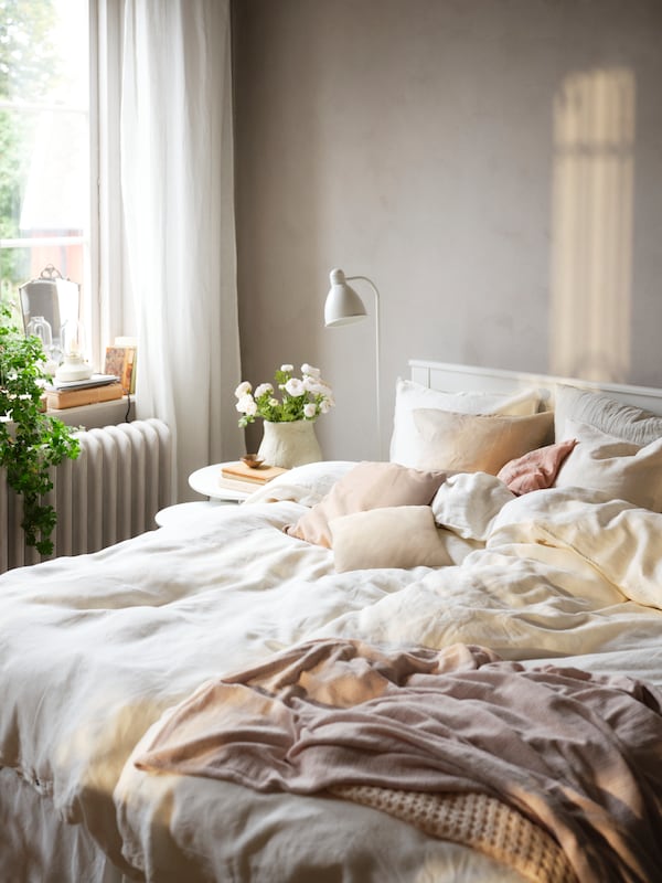 A white SONGESAND bed with white DYTÅG bed linen and other textile products in different colours stands in a sunny bedroom.