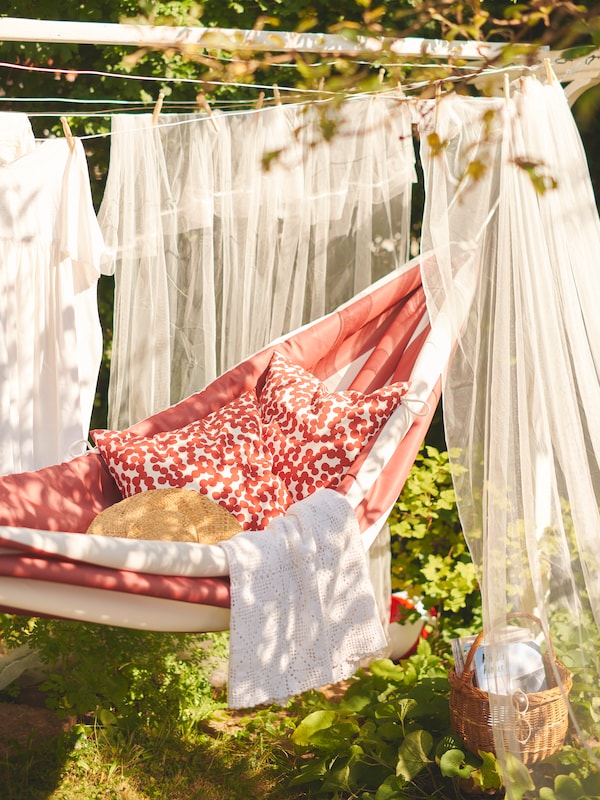 A red and white HAMNÖN hammock with cushions inside it hangs outdoors, with sheer white curtains hanging around it.