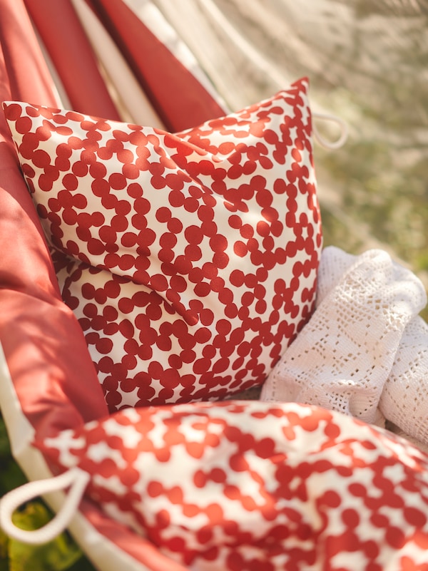 Cushions inside GULLBERGSÖ cushion covers with a red dot pattern lie in a HAMNÖN hammock which is hanging outdoors.