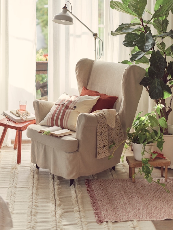 A STRANDMON wing chair sits in a living room, with cushions, including one in a striped VEDMAL cushion cover, lying on it.