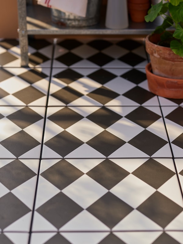 MÄLLSTEN floor decking with a checkerboard pattern covers a balcony floor. A HYLLIS shelf unit and a plant are set on it.