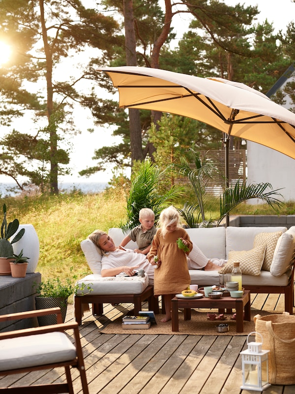 A woman relaxes on an ÄPPLARÖ modular sofa with her children. A SEGLARÖ/SVARTÖ hanging umbrella with base provides shade.