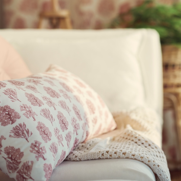 A white/pink VATTENMÅRA cushion sits on a piece of fabric in a corner of a white BACKSÄLEN 3-seat sofa.