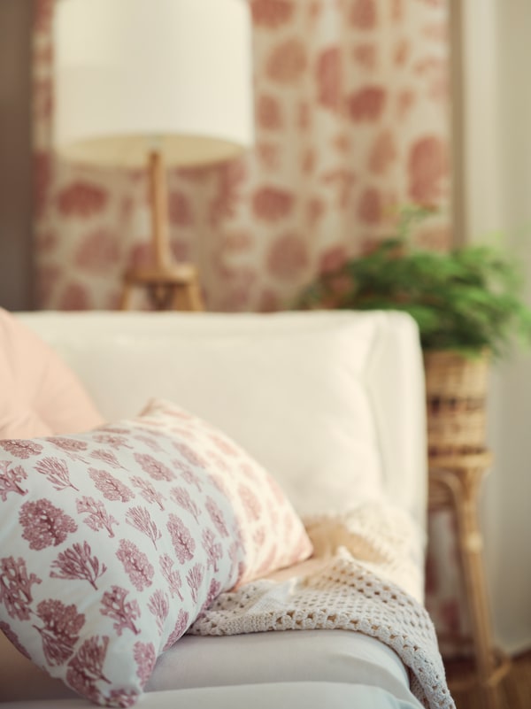 A white/pink VATTENMÅRA cushion sits on a piece of fabric in a corner of a white BACKSÄLEN 3-seat sofa.