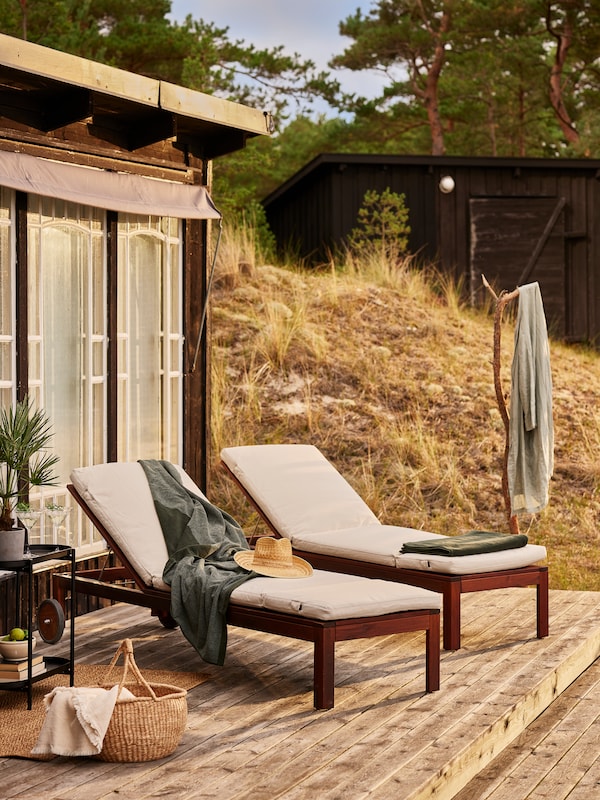 Two ÄPPLARÖ sun loungers on a deck beside a rocky hill, with bath sheets and a hat on them.