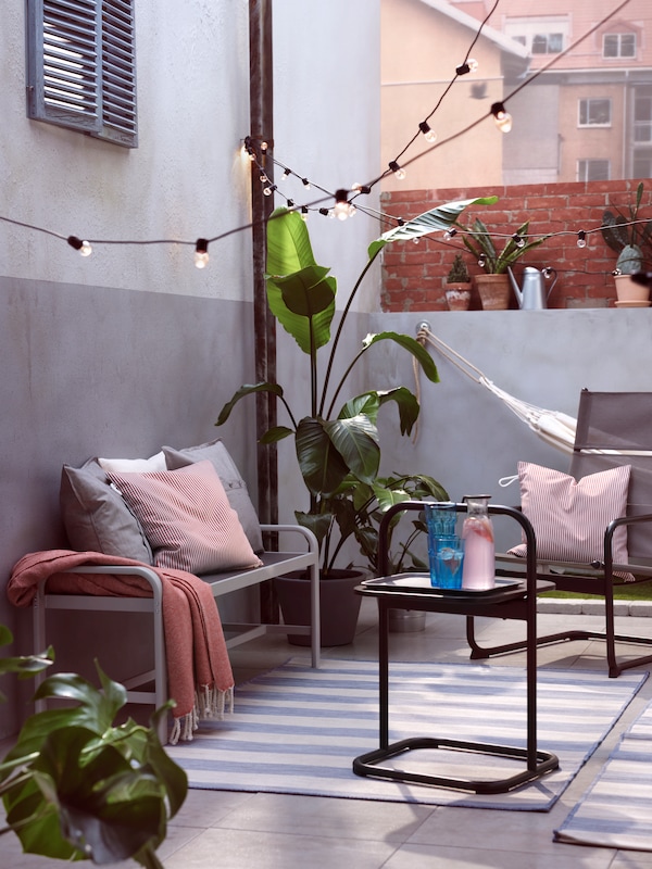 A HUSARÖ outdoor armchair and side table, UTSUND lighting chains, GULLBERGSÖ cushions and SJÄLLAND bench in a communal yard.