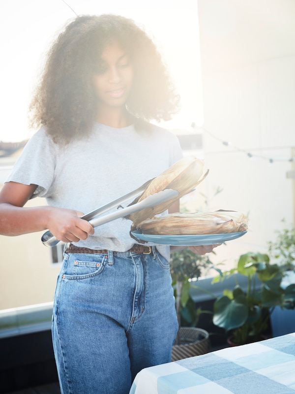 A woman uses GRILLTIDER tongs to lift a grilled corn cob from a FÄRGKLAR serving plate. A RUTIG tablecloth covers the table.