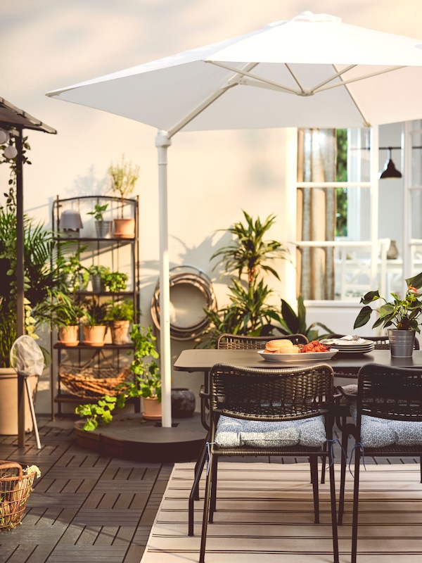 A white HÖGÖN umbrella on a sunny garden terrace with lots of plants, over a dark grey VIHOLMEN table and LÄCKÖ chairs.