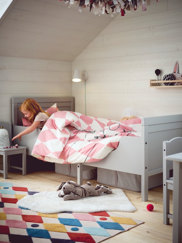 A girl in a grey SUNDVIK extendable bed with pink and white BUSENKEL bed linen reaching for a toy on a chair beside the bed.