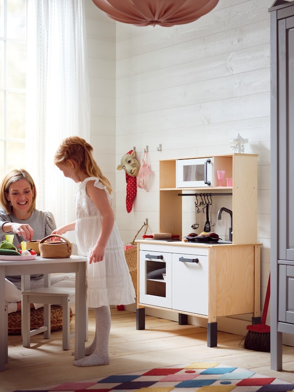 A girl standing at a children’s table playing with a DUKTIG fruit basket and other toys with her mother, by a play kitchen.