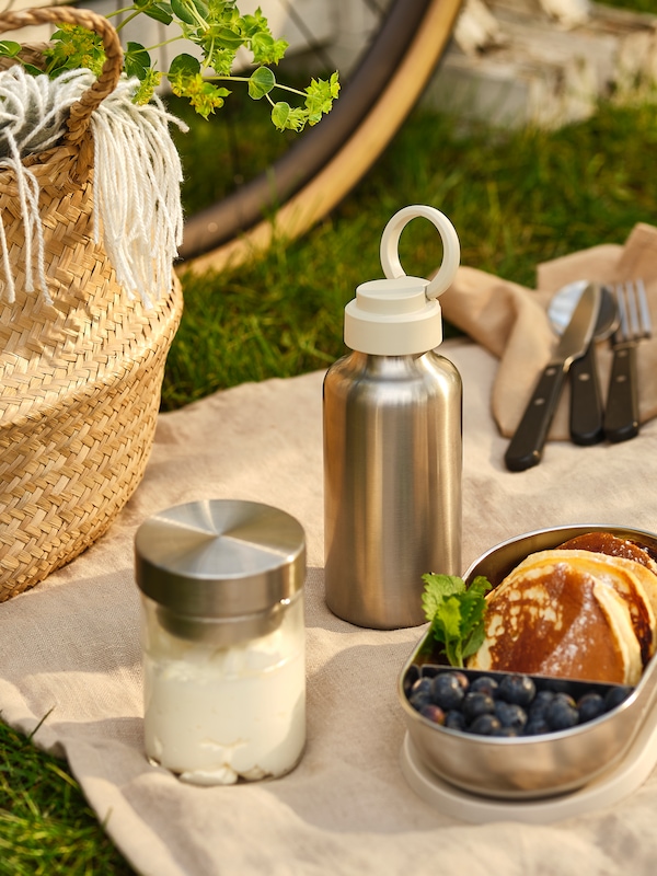 An ENKELSPÅRIG stainless steel water bottle, a HALVVARM food container and picnic accessories laid out on a blanket.