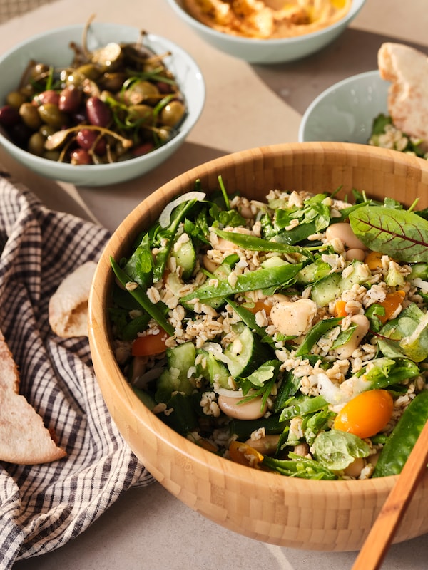 A tabletop with a bamboo BLANDA MATT serving bowl filled with salad, placed next to side dishes in smaller, matt green bowls.