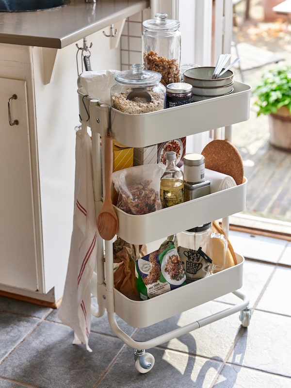 A kitchen with a white RÅSKOG cart filled with VARDAGEN glass jars, GLADELIG bowls, food, and accessories.