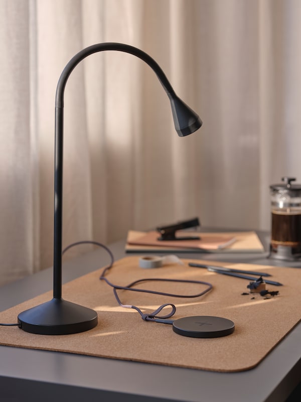 A desktop with a dark-grey LILLHULT USB-A-to-USB-C cord, a black NÄVLINGE LED work lamp and a black LIVBOJ wireless charger.