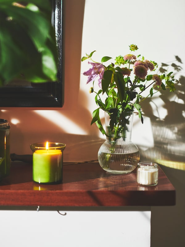 A lit HEDERSAM fresh-grass scented candle and a PÅDRAG glass vase with flowers placed on a mantlepiece.
