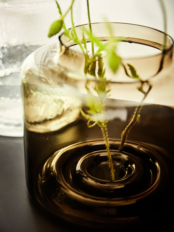 The stems of two plants stand in a green-brown KONSTFULL vase which is standing on a dark surface.