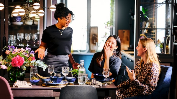 Three laughing women around a dining table laid with ENTUSIASM side plates, SÄLLSKAPLIG wine glasses and a carafe.