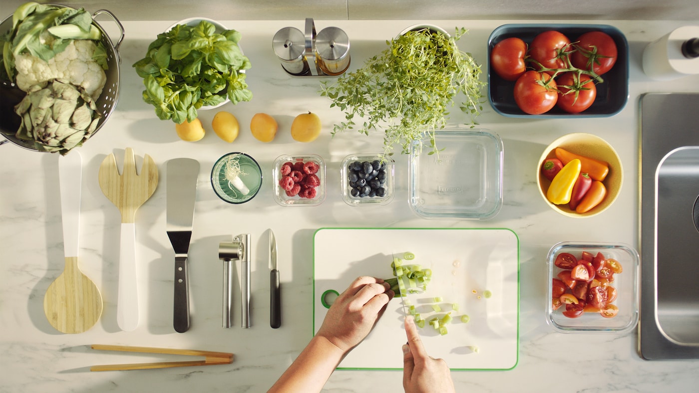 Two hands slice a spring onion on a green/white MATLUST chopping board, next to utensils, vegetables, fruits and herbs.