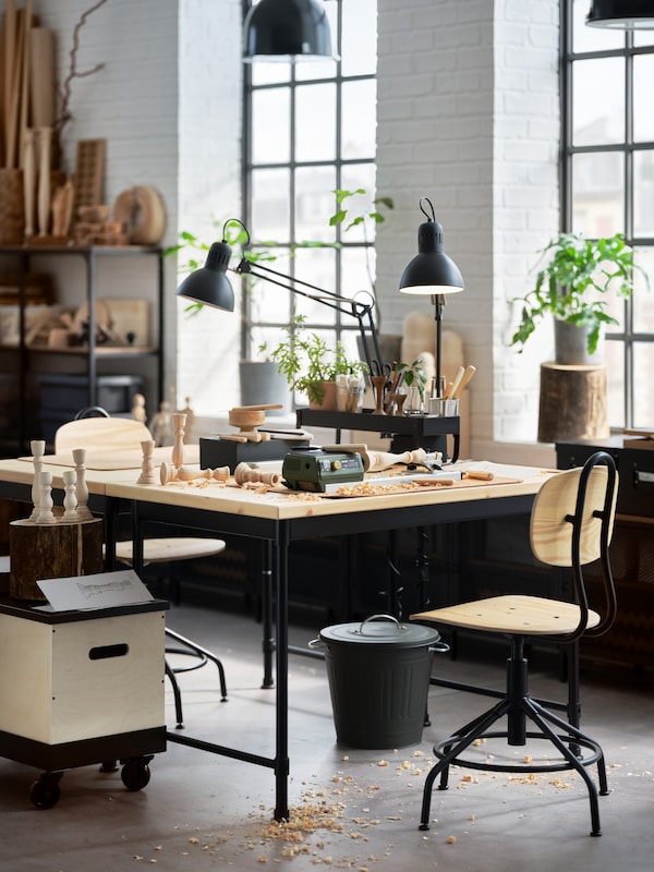 Two pine desks, two pine/black swivel chairs, two dark grey work lamps, big windows and potted plants.