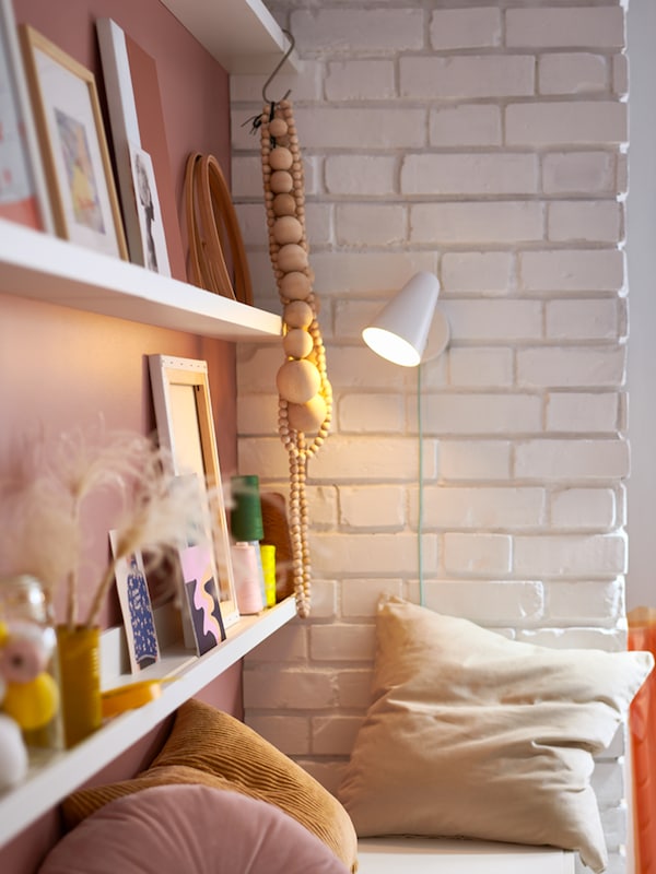 Two rows of white MOSSLANDA picture ledges, framed pictures, accessories, a wall lamp and a white bench with cushions.