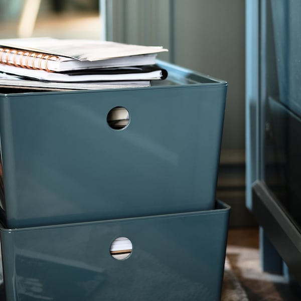 Two stacked, turquoise KUGGIS storage boxes holding notebooks and paper on a rug by a BILLY glass-door bookcase.