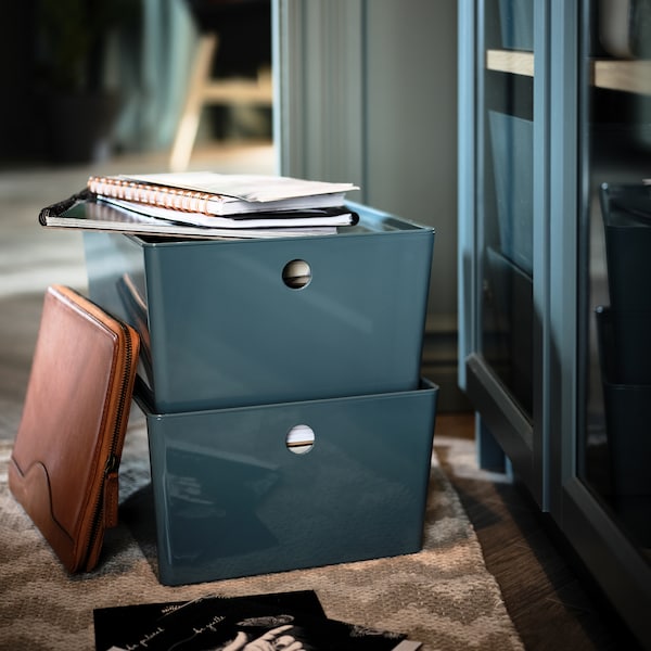 Two stacked, turquoise KUGGIS storage boxes holding notebooks and paper on a rug by a BILLY glass-door bookcase.