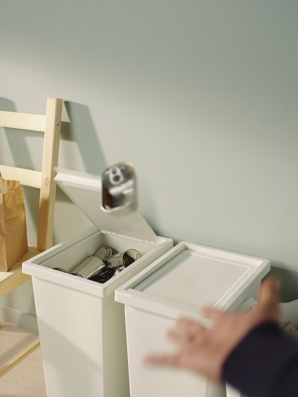 Two, white, HÅLLBAR recycling bins, the lid of one open as a hand in the foreground tosses in a can.