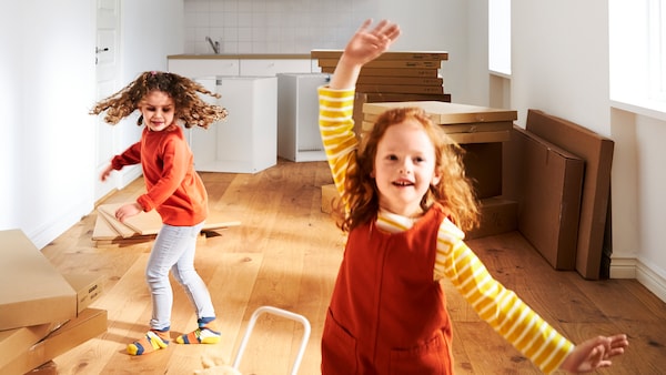 Two young girls in a bare room with wooden floorboards and white walls, in front of piles of IKEA flat-packed products.